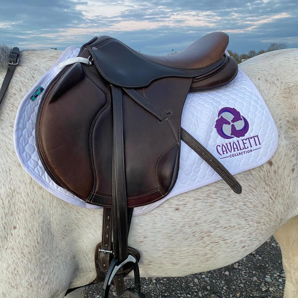 Close‑up of a brown saddle on a white quilted Cavaletti Collection saddle pad in an outdoor setting.