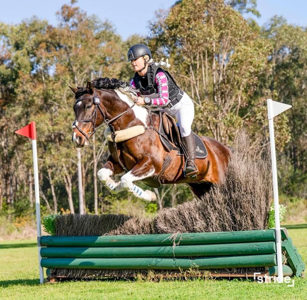 Horse and rider mid‑air over a green brush cross‑country obstacle in an outdoor field. Photo credit: millybeventing.
