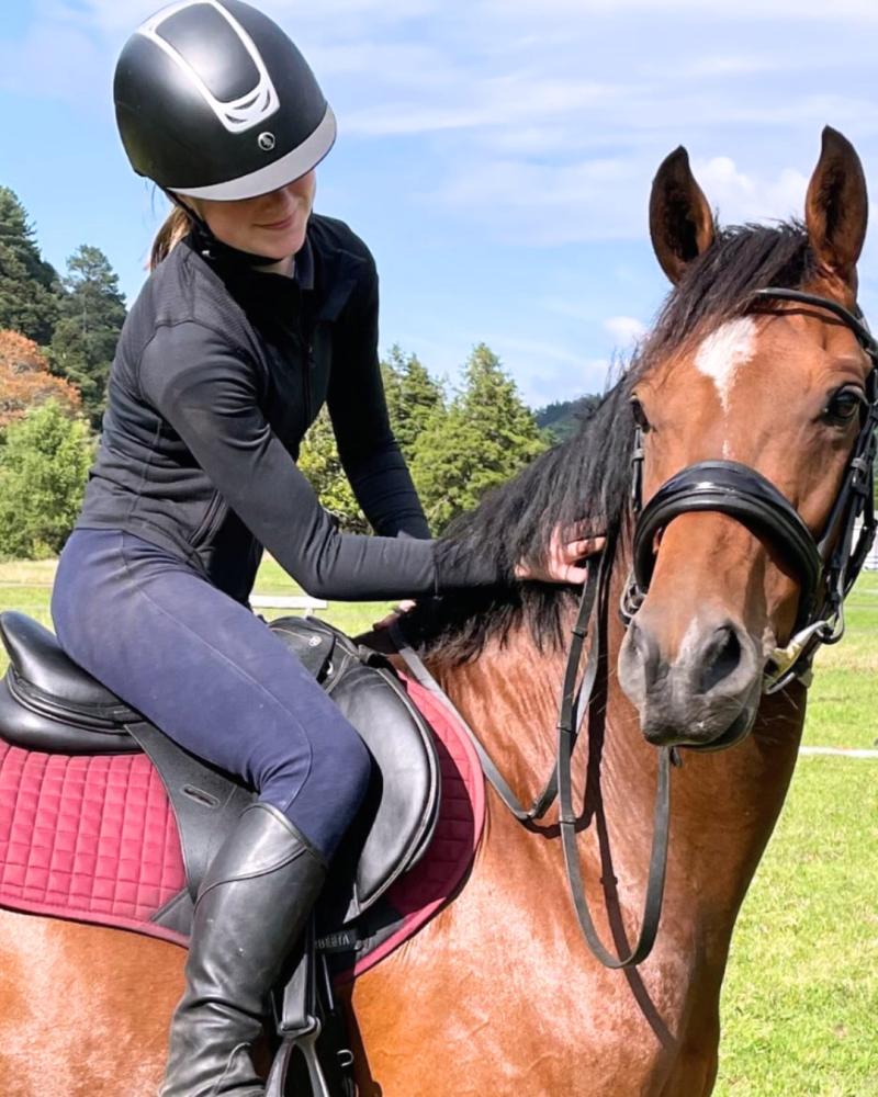 Rider sitting on a brown horse with a maroon saddle pad in an outdoor grassy setting.